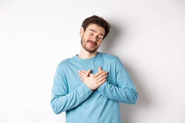 Handsome young man feeling romantic, holding hands on heart and thinking about good pleasant memories, standing over white background