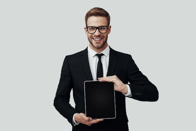 Handsome young man in formalwear showing copy space on digital tablet while standing against grey background