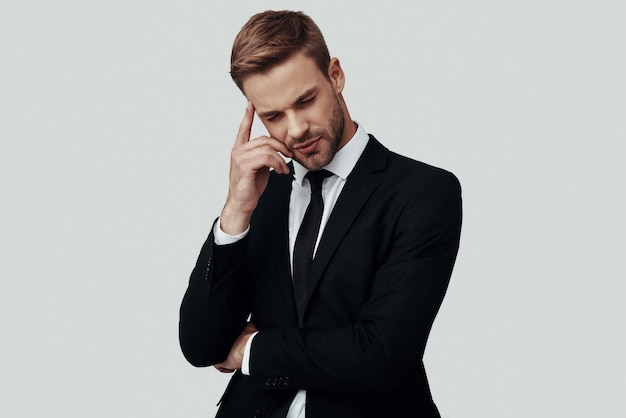 Handsome young man in formalwear thinking about something while standing against grey background