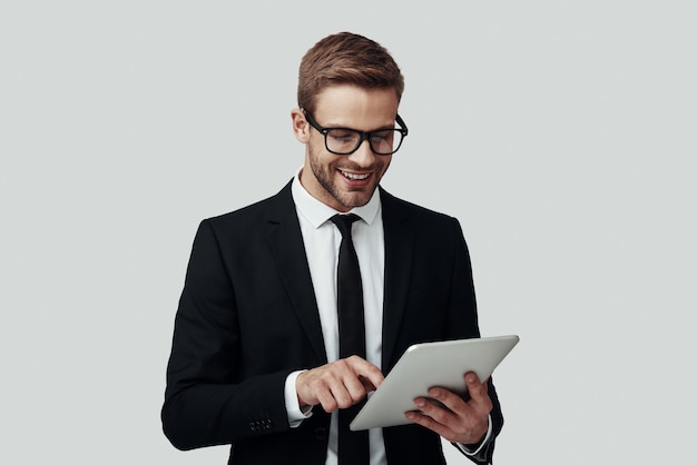 Handsome young man in formalwear working using digital tablet while standing against grey background