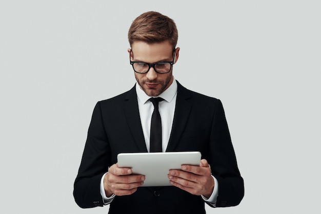 Handsome young man in formalwear working using digital tablet while standing against grey background