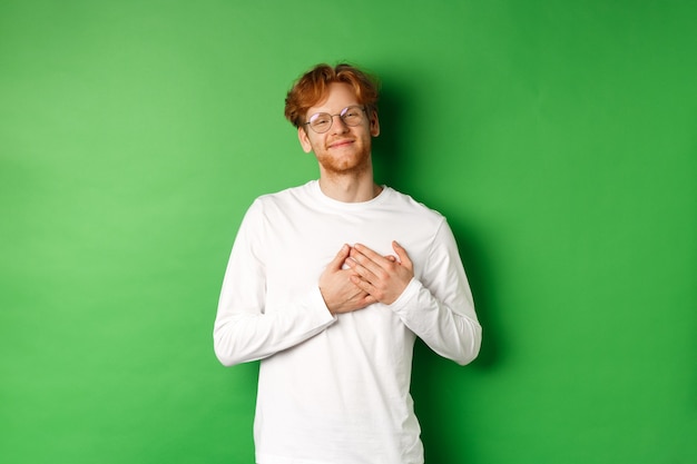 Handsome young man in glasses and white long-sleeve smiling, holding hands on heart with grateful face, thanking you, standing over green background