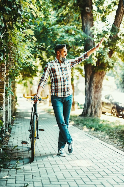 ハンサムな若い男が自転車を持って街に出かけ、その横に立って誰かに手を振っています。