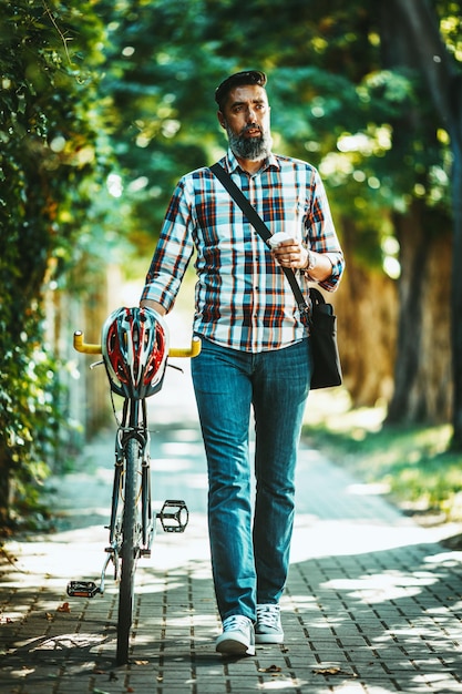 ハンサムな若い男が自転車を持って街に出かけ、コーヒーを片手に歩いてどこへ行くのか考えています。