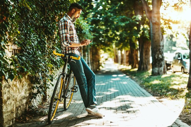 A handsome young man goes to the city with his bike, standing beside it, waiting for someone and reading text message.