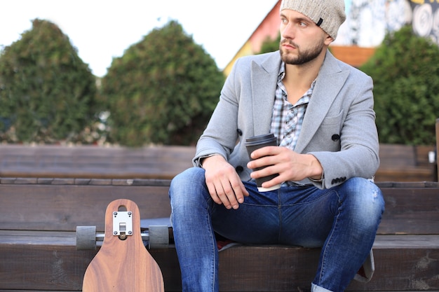 Handsome young man in grey coat and hat, resting, sitting with longboard drinking coffee. Urban skateboarding concept.