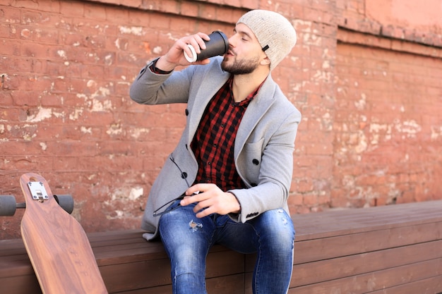 Handsome young man in grey coat and hat, resting, sitting with\
longboard drinking coffee. urban skateboarding concept.