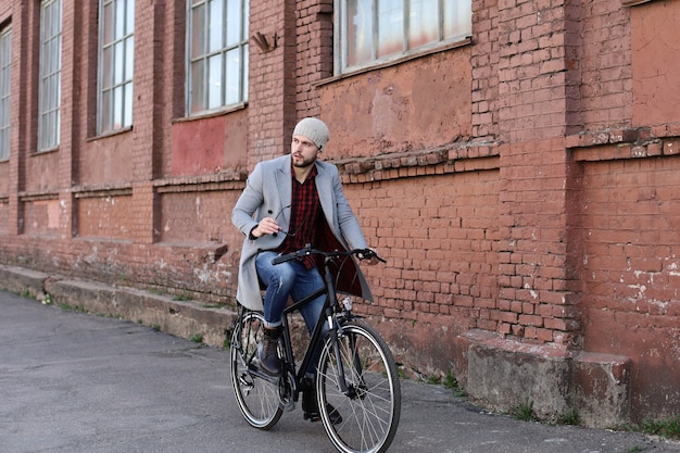회색 코트와 모자를 쓴 잘생긴 청년이 도시의 자전거 거리를 타고 있습니다.