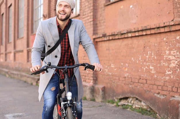 街の自転車通りに乗って灰色のコートと帽子のハンサムな若い男。