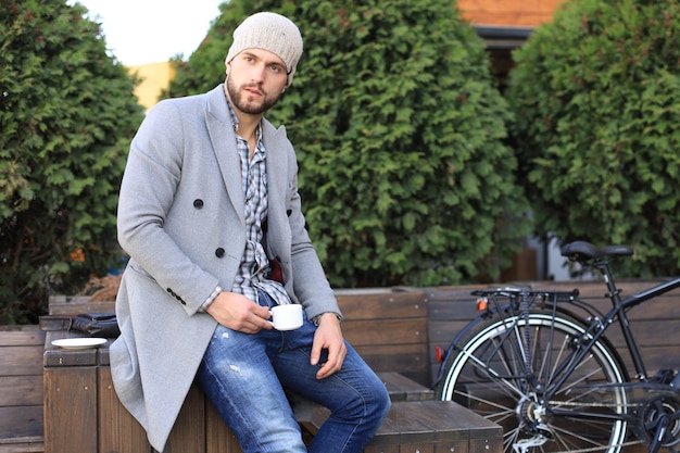 Handsome young man in grey coat and hat sitting on a bench relaxed drinking coffee and thinking near his bicycle.