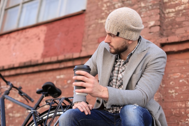 灰色のコートと帽子をかぶったハンサムな若い男がベンチに座って、コーヒーを飲みながら自転車の近くで考えてリラックスしました。