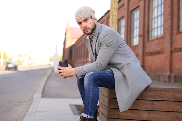 Handsome young man in grey coat and hat sitting on a bench relaxed drinking coffee.