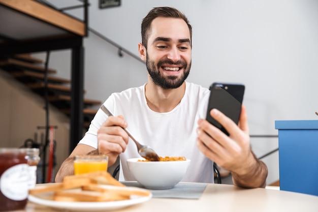 Handsome young man having breakfast while sitting at the kitchen, holding mobile phone