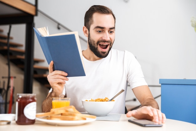 Handsome young man having breakfast while sitting at the kitchen, reading a book