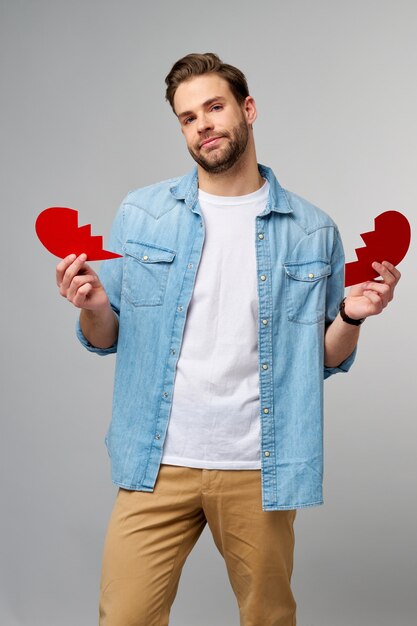 handsome young man holding broken paper red valentine heart standing over grey