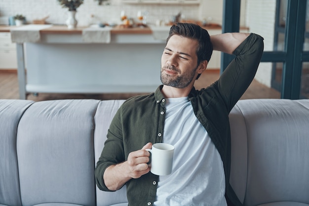 Handsome young man holding cup with drink while sitting on the sofa at home