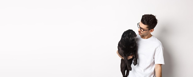 Handsome young man holding his black pug dot and pet owner looking each other faces standing against