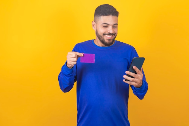 Handsome young man holds credit card and smartphone isolated on yellow background Online shopping Online payment