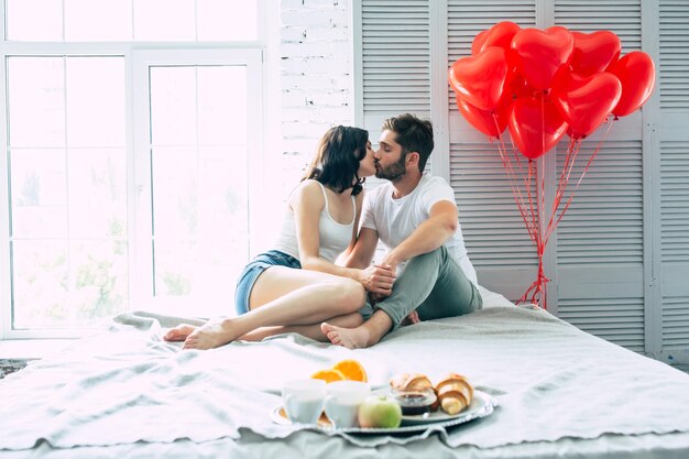 Красивый молодой человек стоит с романтическим завтраком на подносе, он приготовил его для своей красивой улыбающейся милой девушки или жены, которая лежит на кровати и ждет его
