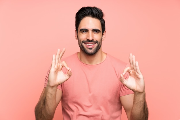 Красивый молодой человек над изолированной розовой стеной показывая одобренный знак с пальцами