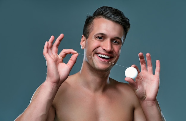 Handsome young man isolated. portrait of shirtless muscular man is standing on grey background with dental floss in hands. men care concept