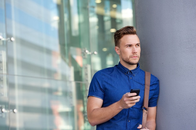 Giovane bello che si appoggia contro la parete fuori con il cellulare