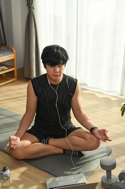 Handsome young man listening to music in earphones and practicing yoga on yoga mat at home.