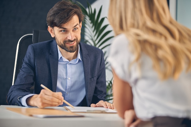 Handsome young man looking at female business partner and smiling while sitting at the table and pointing at clipboard