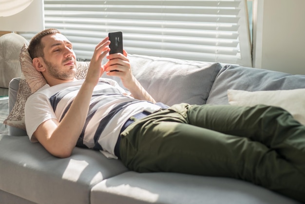 Красивый молодой человек смотрит на свой смартфон, лежа дома на диване