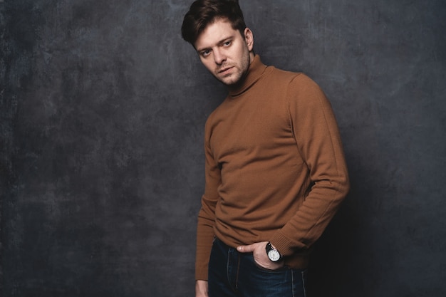 Handsome young man looking. Portrait of young man with hands in pockets leaning against gray wall.