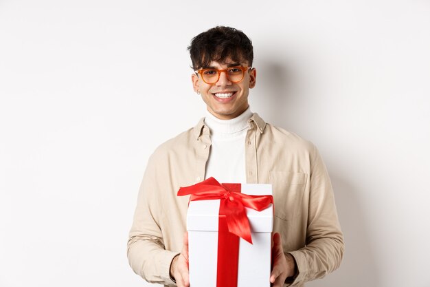 Handsome young man making a gift, holding box with present and smiling , standing on white wall.