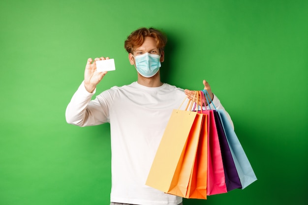 Photo handsome young man in medical mask, showing plastic credit card and shopping bags with items purchased with discount, green background.