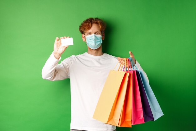 Handsome young man in medical mask, showing plastic credit card and shopping bags with items purchased with discount, green background