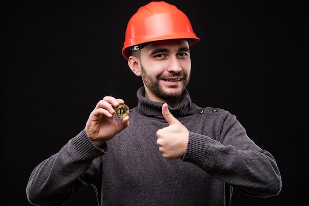 Handsome young man miner in protective hemlet pointed bitcoin with thumbs up isolated on black