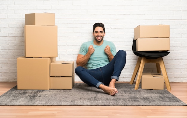 Handsome young man moving in new home among boxes celebrating a victory