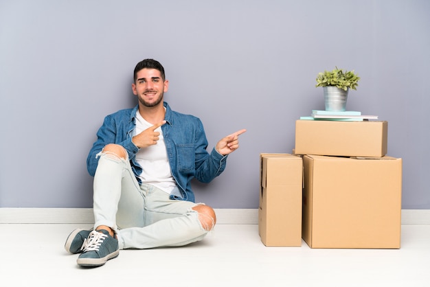 Handsome young man moving in new home among boxes pointing finger to the side