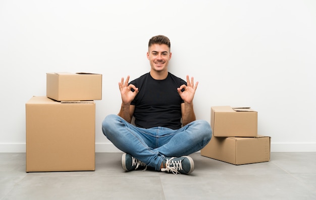 Handsome young man moving in new home among boxes showing an ok sign with fingers