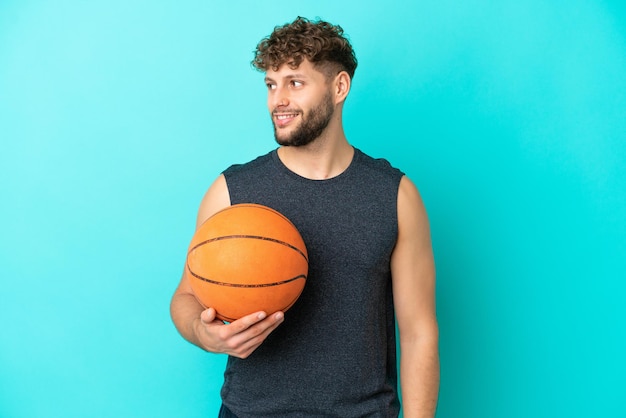 Bel giovane uomo che gioca a basket isolato su sfondo blu guardando al lato e sorridente