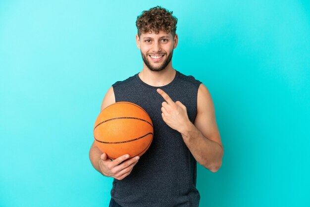 Bel giovane che gioca a basket isolato su sfondo blu che punta al lato per presentare un prodotto