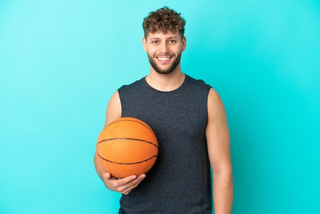 Bel giovane uomo che gioca a basket isolato su sfondo blu in posa con le braccia all'anca e sorridente
