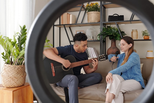 Handsome young man playing guitar when his girlfriend is singing view through the ring light