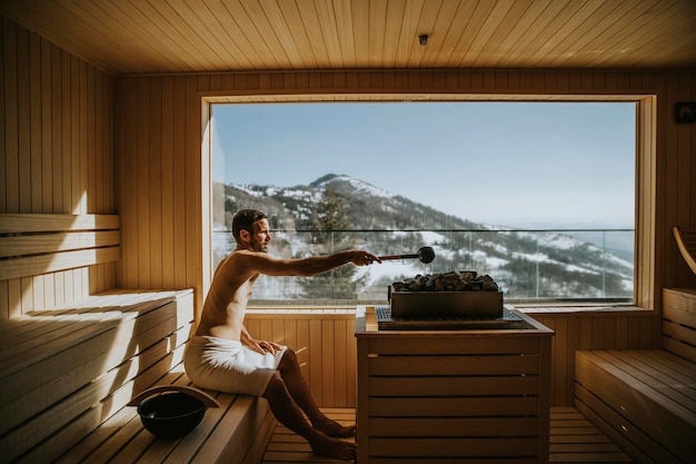 Handsome young man pouring water onto hot stone in the sauna