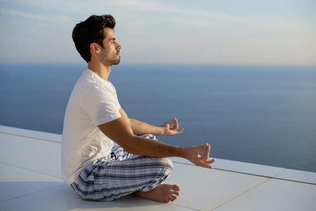 handsome young man practicing yoga on in modern home terace with ocean and sunset in background