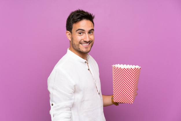 Handsome young man over purple wall holding popcorns