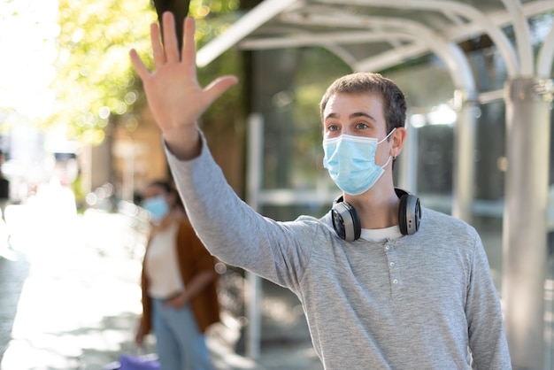 A handsome young man raising his hand to stop the bus Boy at the bus station wearing a face mask during Coronavirus pandemic