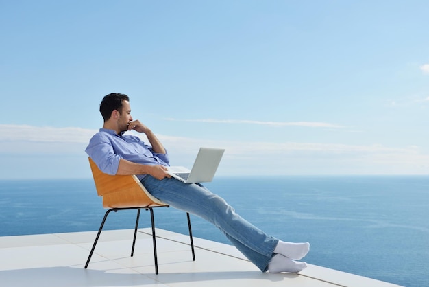 handsome young man relaxing and working on laptop computer at home balcony while looking sunset