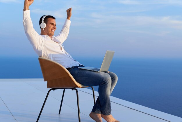 handsome young man relaxing and working on laptop computer at home balcony while looking sunset