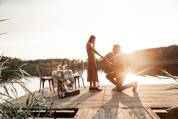 Фото Красивый молодой человек просит руки любимой красивой подруги, прося жениться. парень стоит на коленях во время заката, предлагая обручальное кольцо удивленной женщине на улице у лесного озера