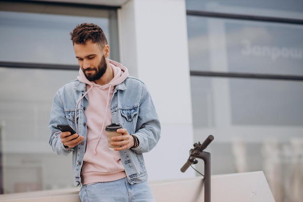 Handsome young man riding electric scooter using phone and drinking coffee