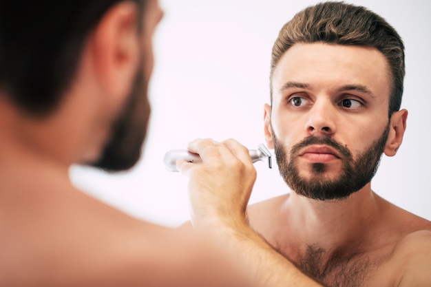 Красивый молодой человек бреет бороду в ванной комнате. Портрет стильного обнаженного бородатого мужчины, разглядывающего свое лицо в домашнем зеркале.
